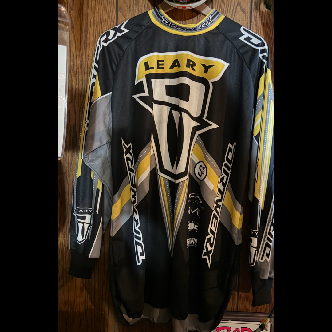 Black, gray, yellow and white DIRTWERX jersey marked LEARY, hanging against the room’s wood-paneled wall.