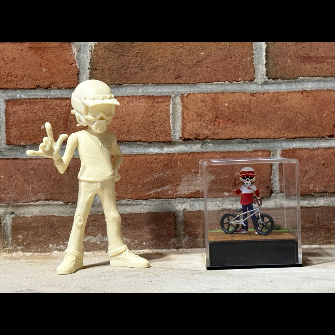 Large unpainted Radical Rick figure beside a small enclosed 1:24-scale Radical Rick bicycle scene against a brick wall.