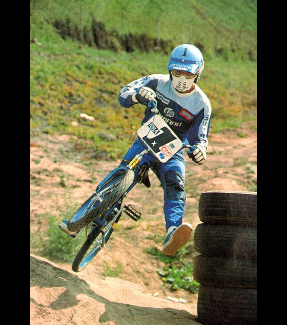 Bobby Encinas airborne in blue-and-gray BMX gear beside stacked tires on an outdoor dirt course