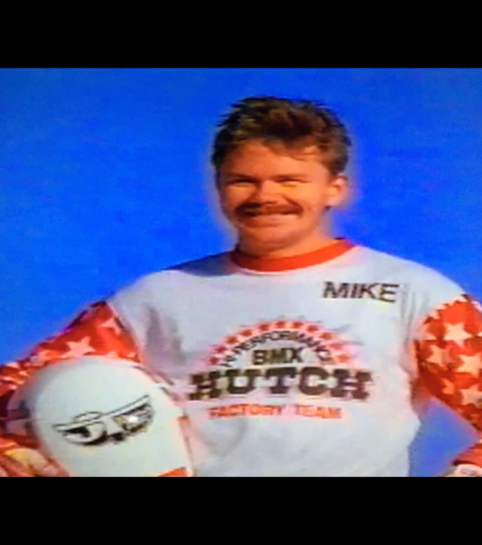 Mustached BMX rider wearing a Hutch Factory Team jersey marked MIKE and holding a full-face helmet