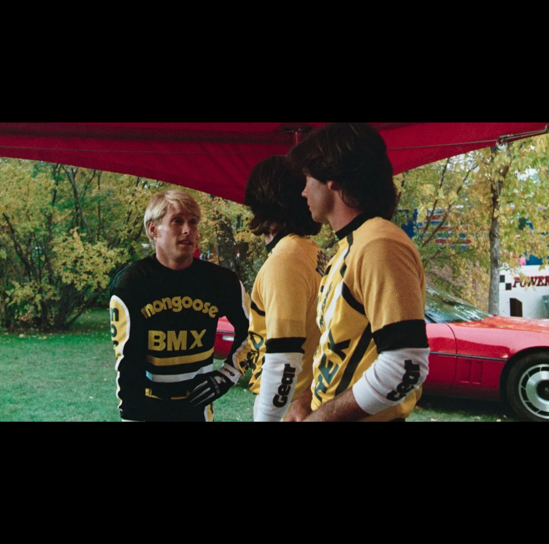 Vintage BMX scene showing riders in Mongoose-branded race jerseys gathered under a red canopy at an outdoor event