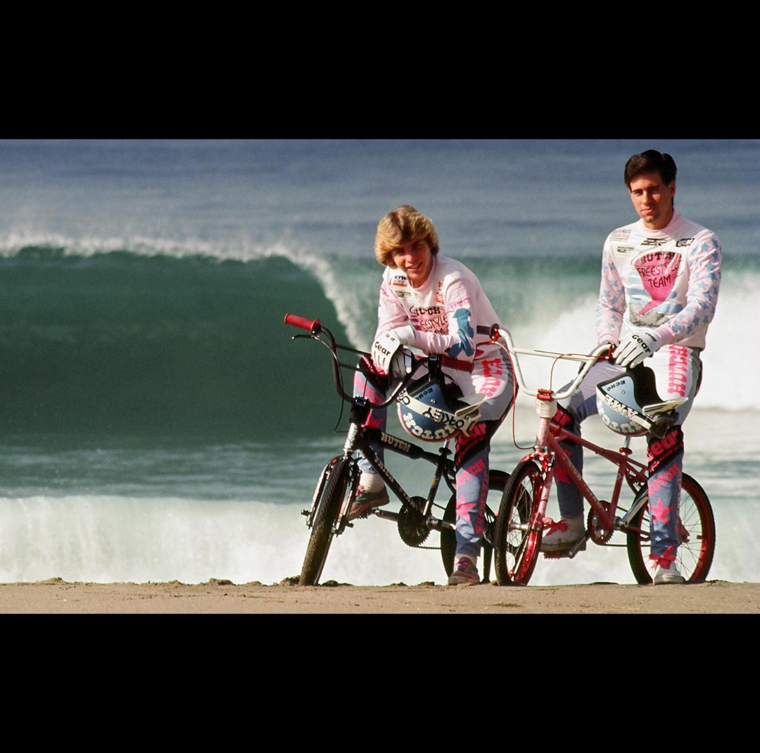 Two Hutch Freestyle Team riders posing with BMX bicycles on a beach as a large ocean wave breaks behind them